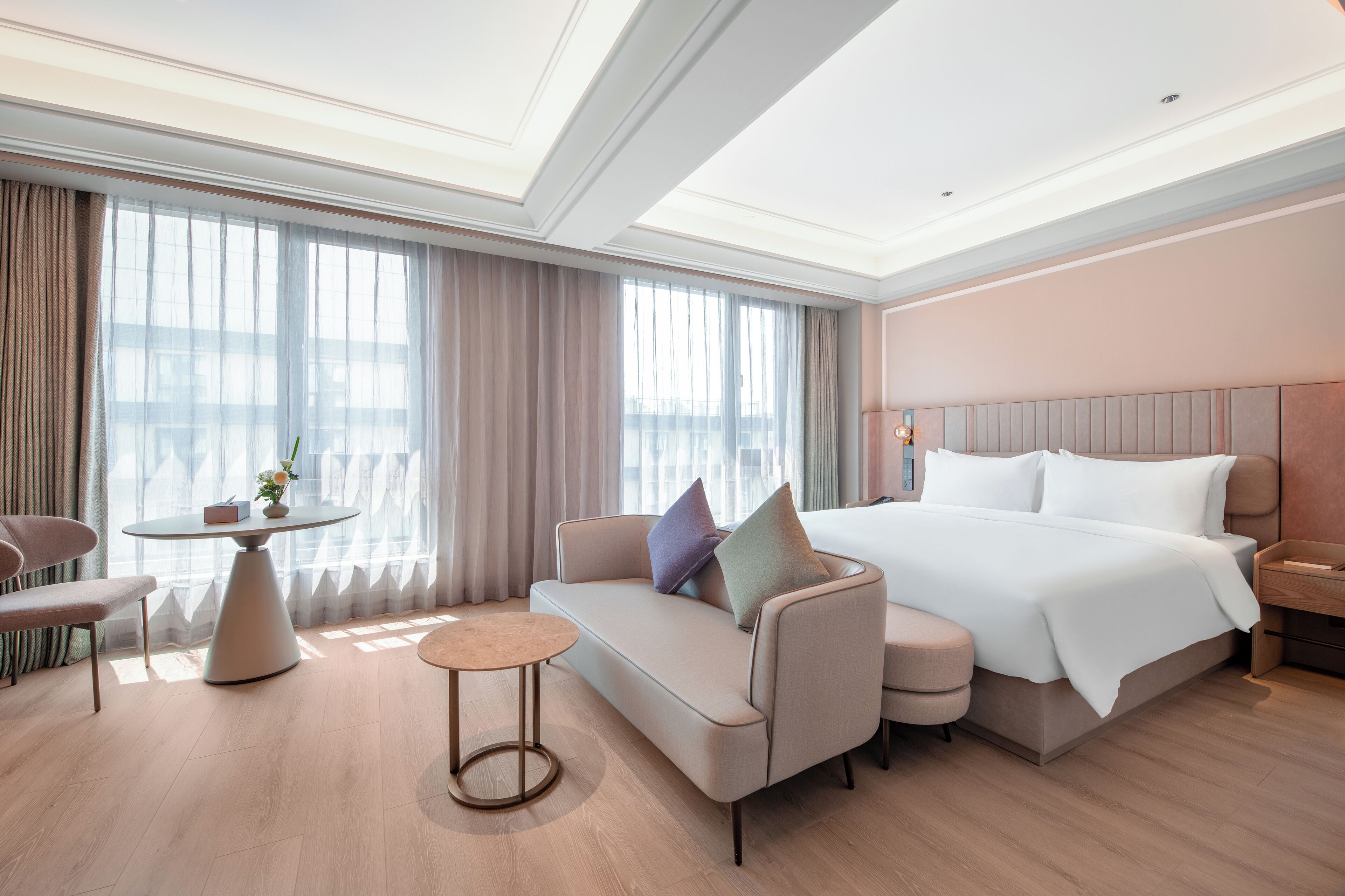 A modern hotel room with a bed, sofa, round table, and a view of the city through large windows.