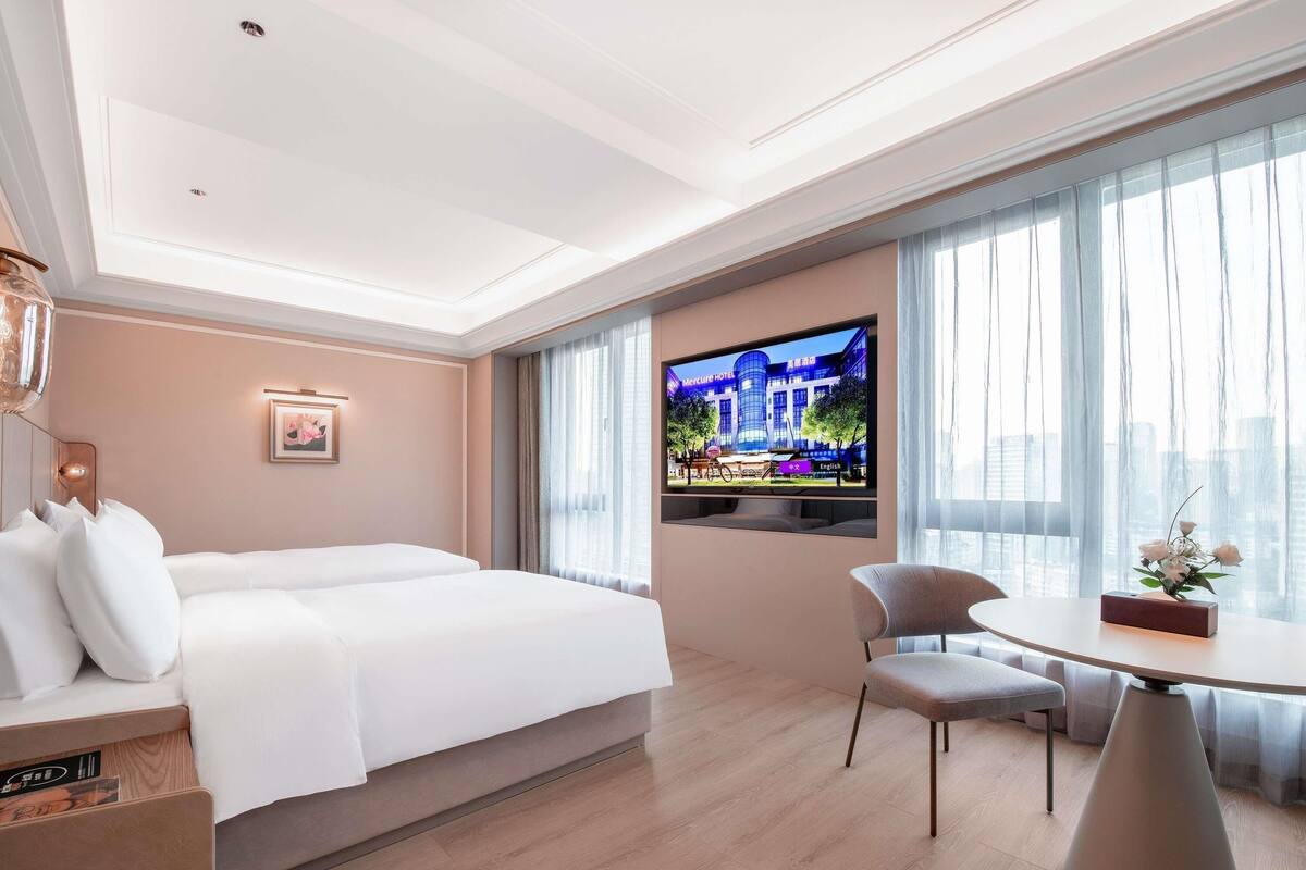 Habitación de hotel con cama, escritorio, silla, televisor montado en la pared y ventana con cortinas.