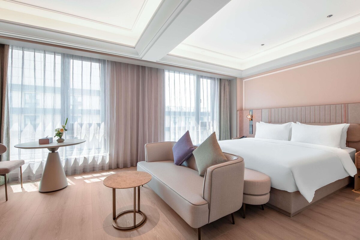 Une chambre d’hôtel avec un lit, un canapé, une table ronde et une vue sur l’extérieur grâce à de grandes fenêtres.