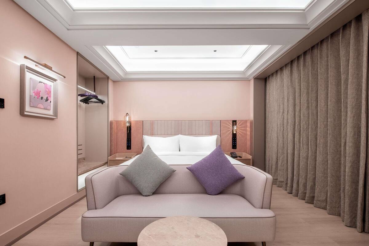 Una habitación de hotel moderna con sofá, una mesita redonda pequeña, una cama con dos almohadas, una mesita de noche, una lámpara de pared y un ventanal grande con cortinas.
