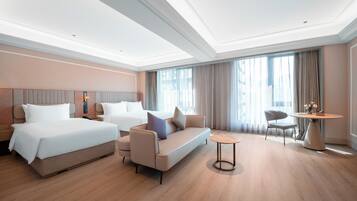 Habitación de hotel con cama, sofá, sillón, mesa redonda y un ventanal con cortinas.