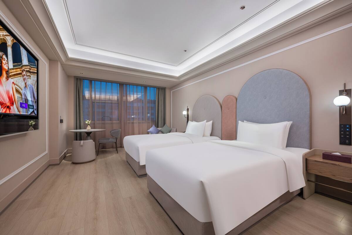 A hotel room with two beds, a TV, a desk, and a large window.
