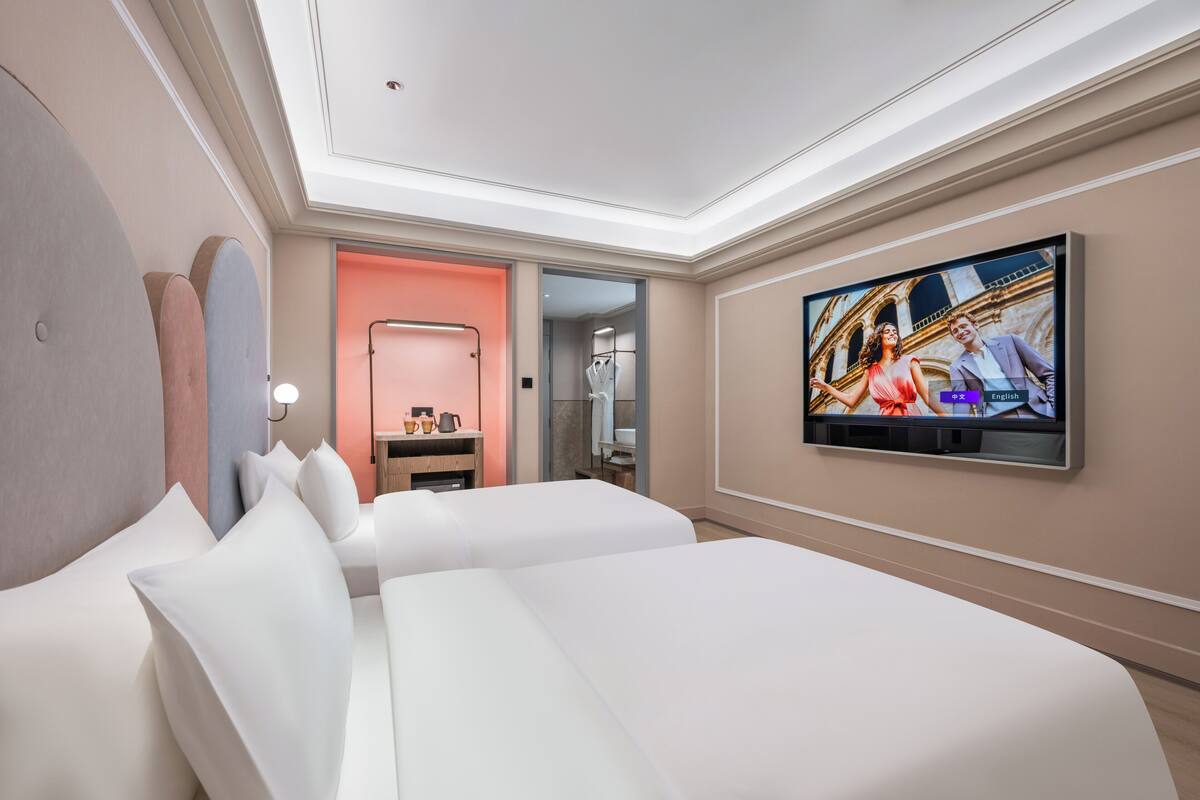 A hotel room with two beds, a TV mounted on the wall, and a view of a building through an open door.