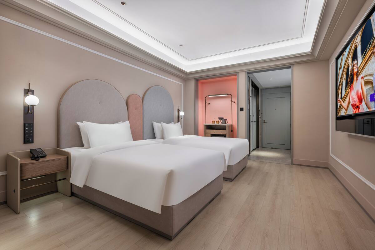 A modern hotel room with two beds, a bedside table with a phone, a TV, and a bathroom visible through an open door.