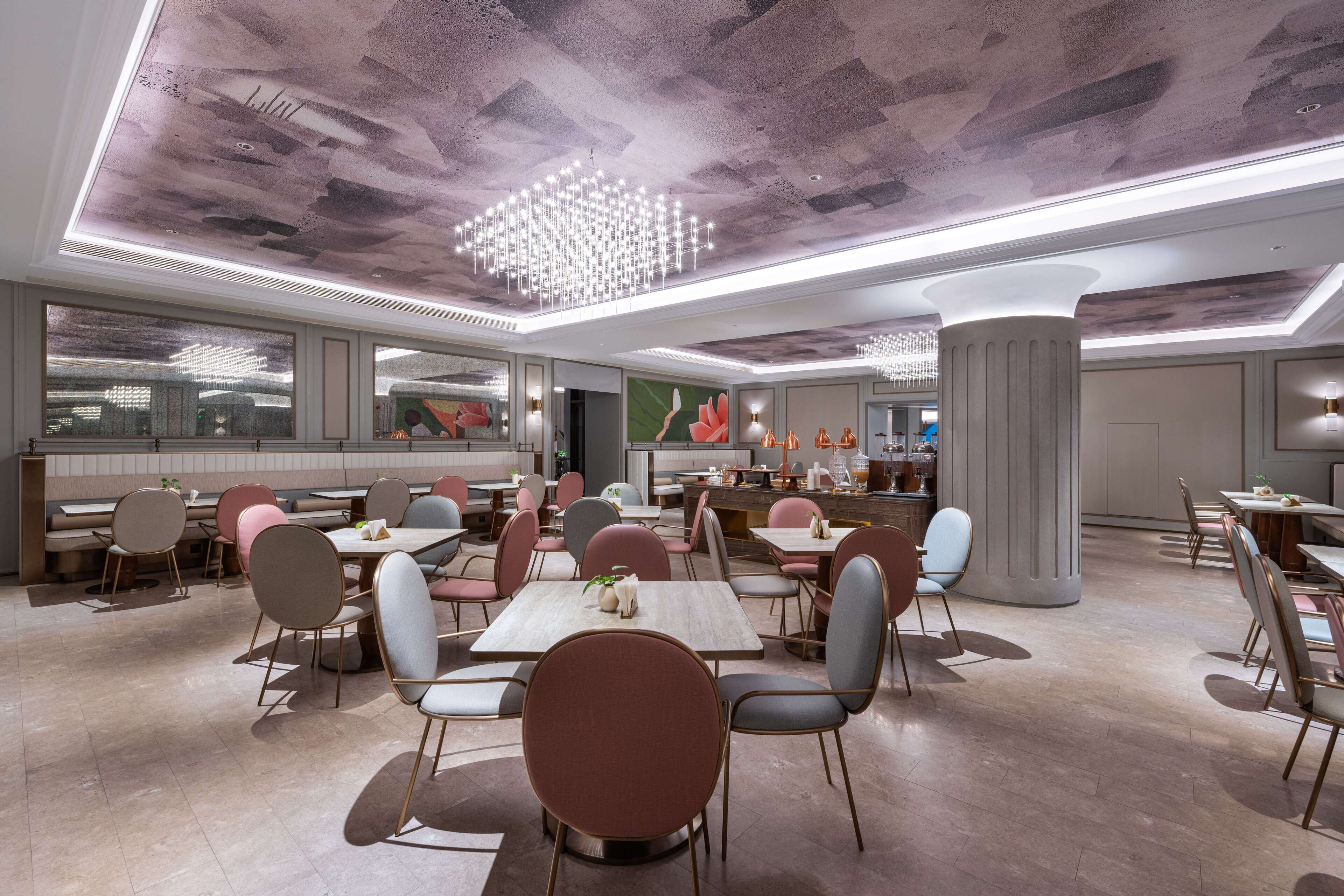 A modern restaurant with a high ceiling, pendant lighting, and a central chandelier. Tables and chairs are arranged in a spacious layout.