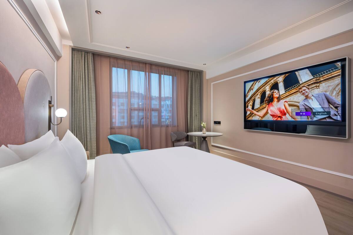 A hotel room with a large bed, a TV mounted on the wall, a small table, and a view of a building through the window.