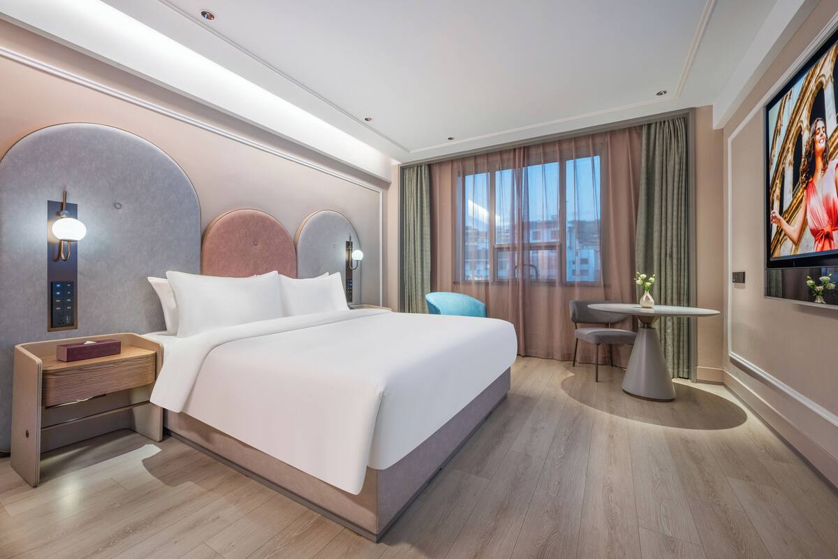 A modern hotel room with a large bed, a bedside table with a lamp, a small round table with a chair, and a TV mounted on the wall.