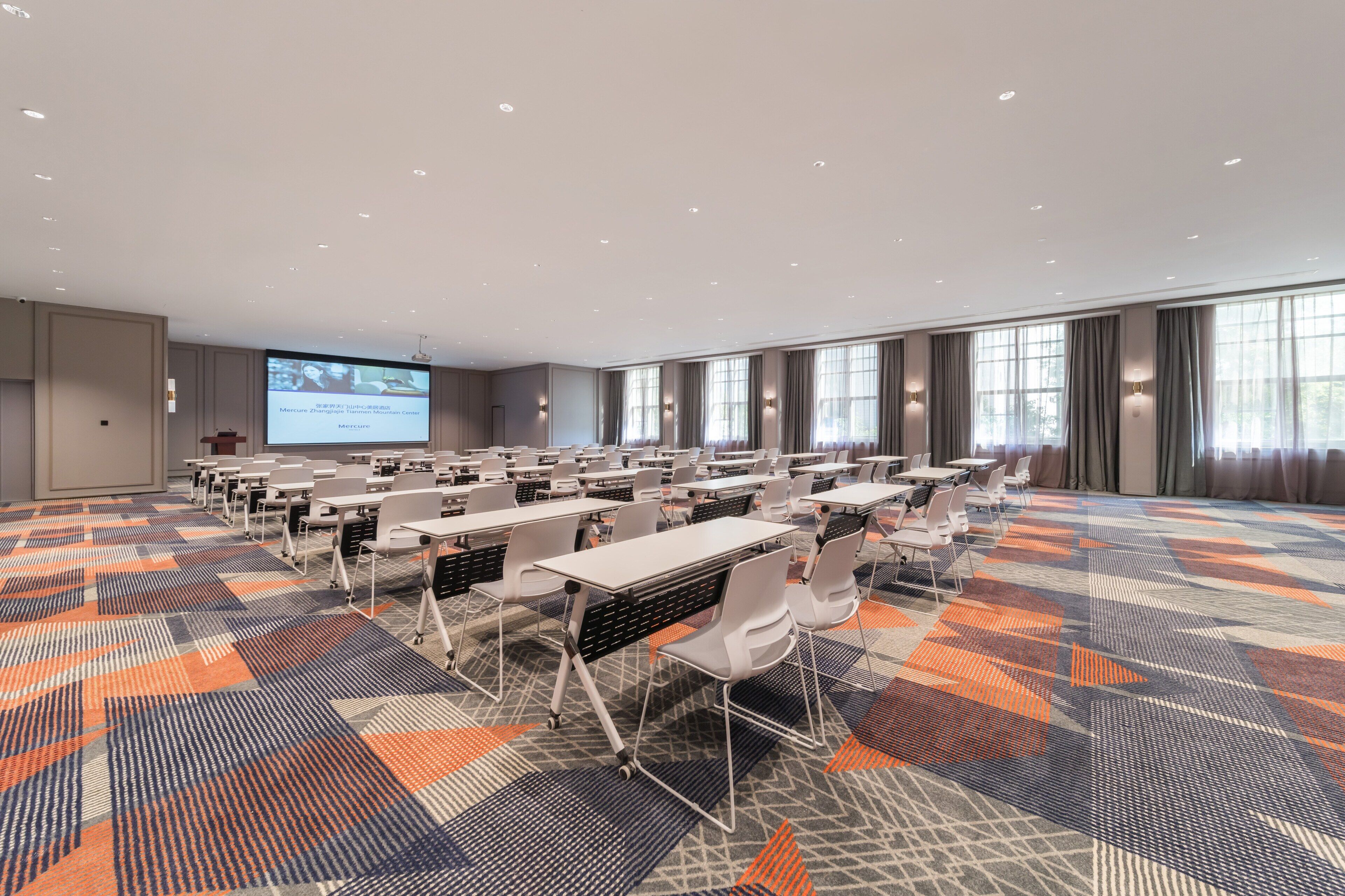 A conference room with rows of tables and chairs, a large screen for presentations, and a patterned carpet.