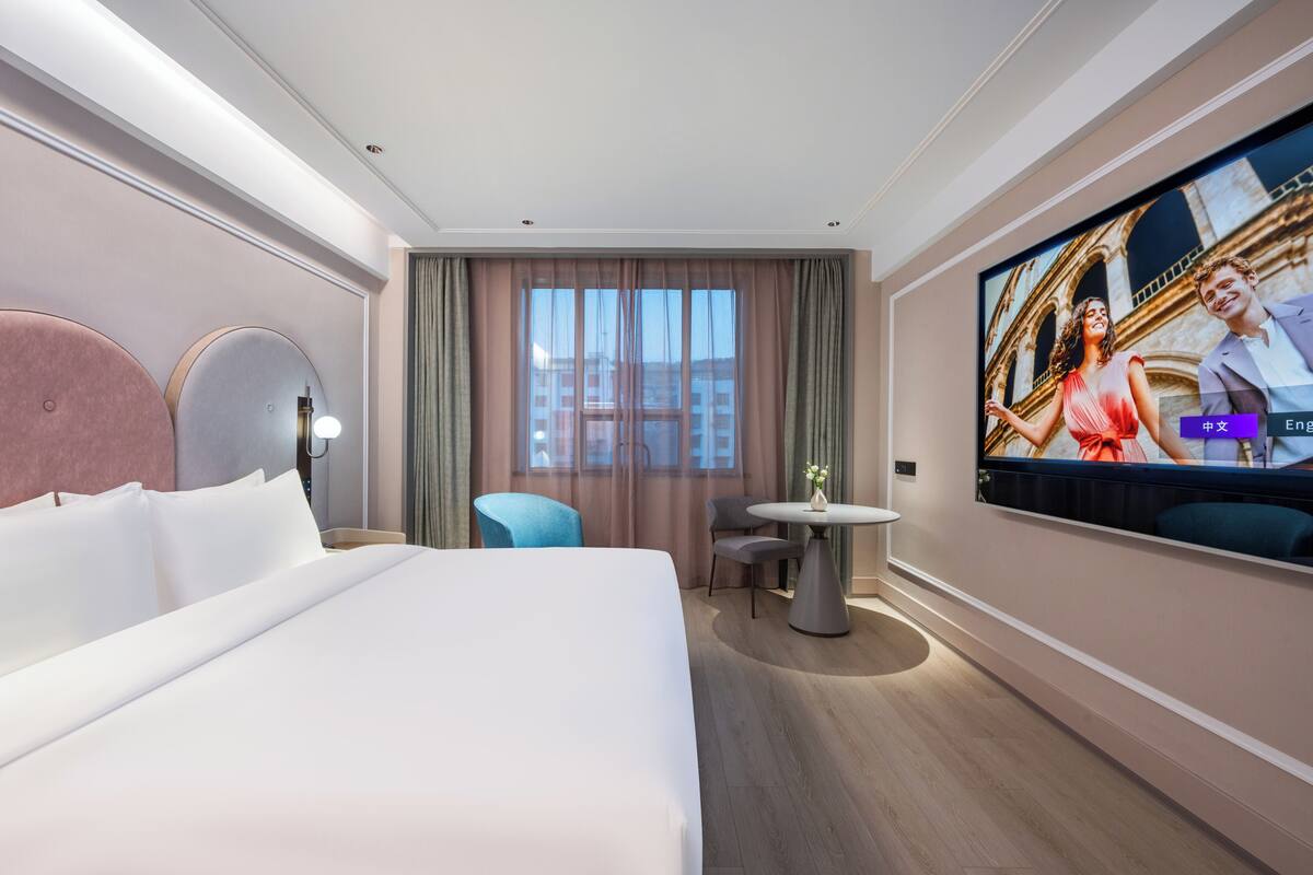 A hotel room with a large bed, a TV mounted on the wall, a small round table, and a window with curtains.