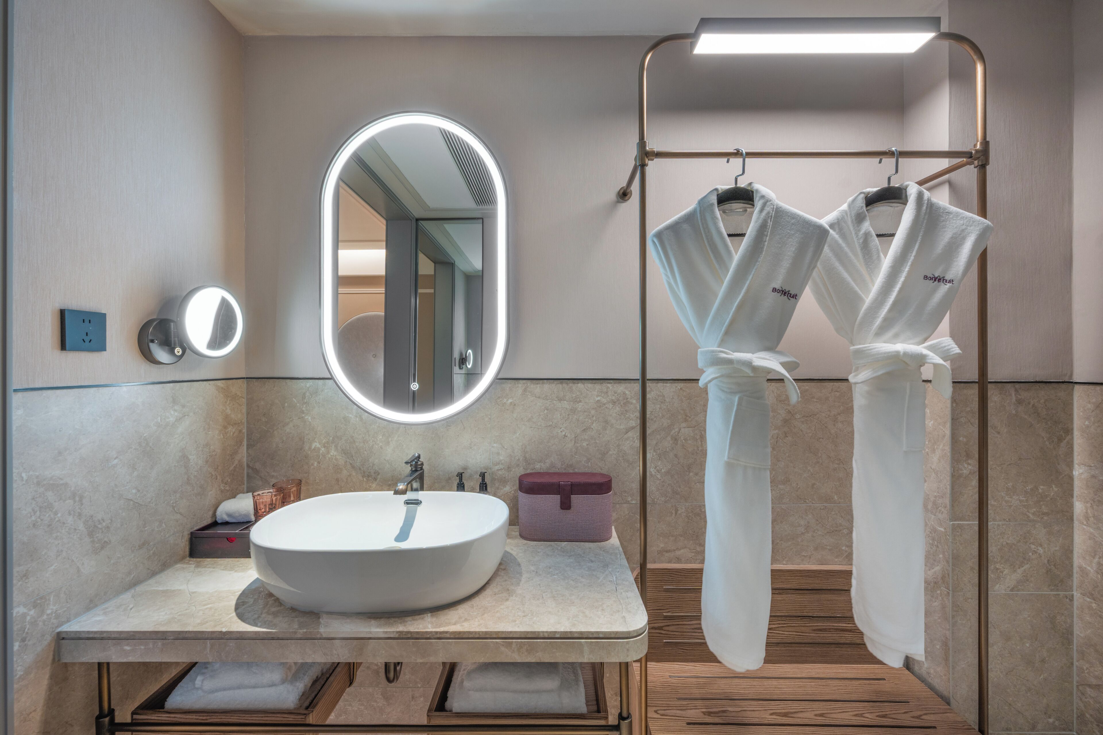 Un baño moderno con un lavamanos redondo, un espejo con iluminación integrada, un perchero con toallas y un mueble de tocador de madera.