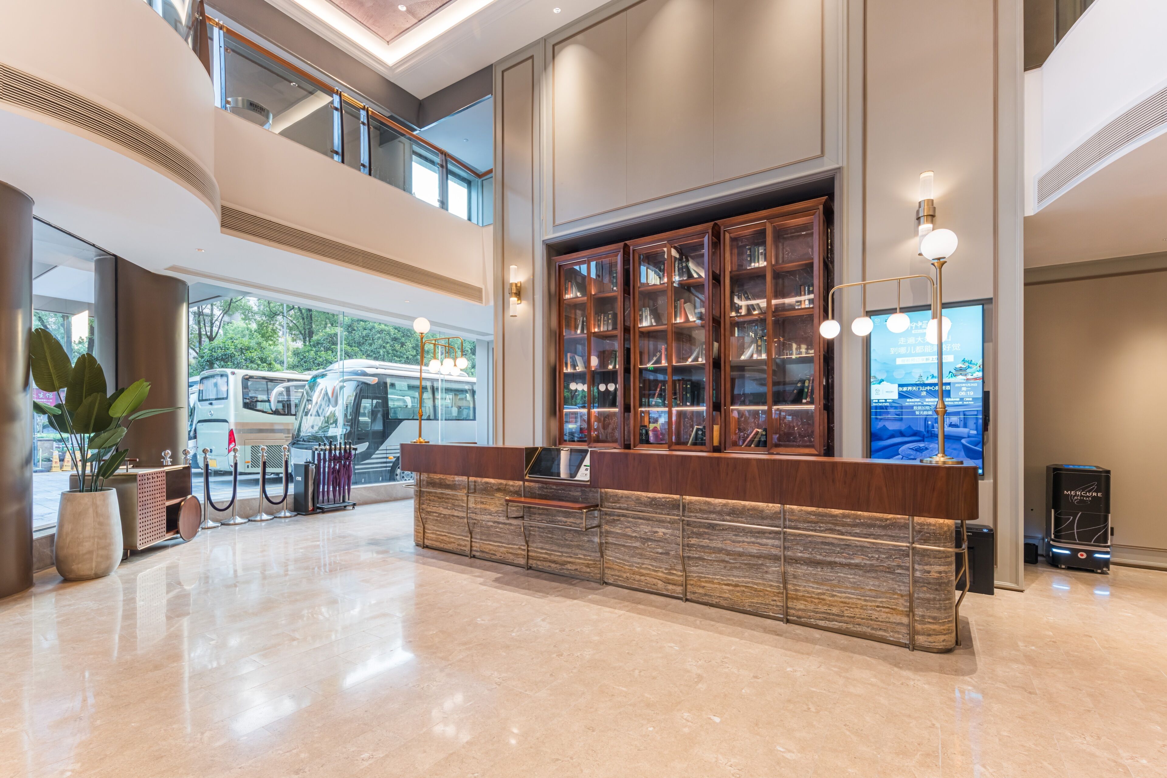 A spacious lobby with a high ceiling, a central reception desk, a large bookshelf, and a modern staircase.