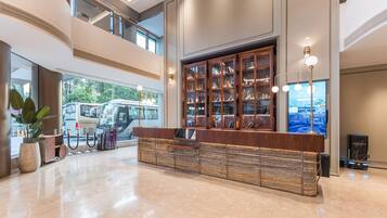 A spacious lobby with a high ceiling, a central reception desk, a large bookshelf, and a modern staircase.