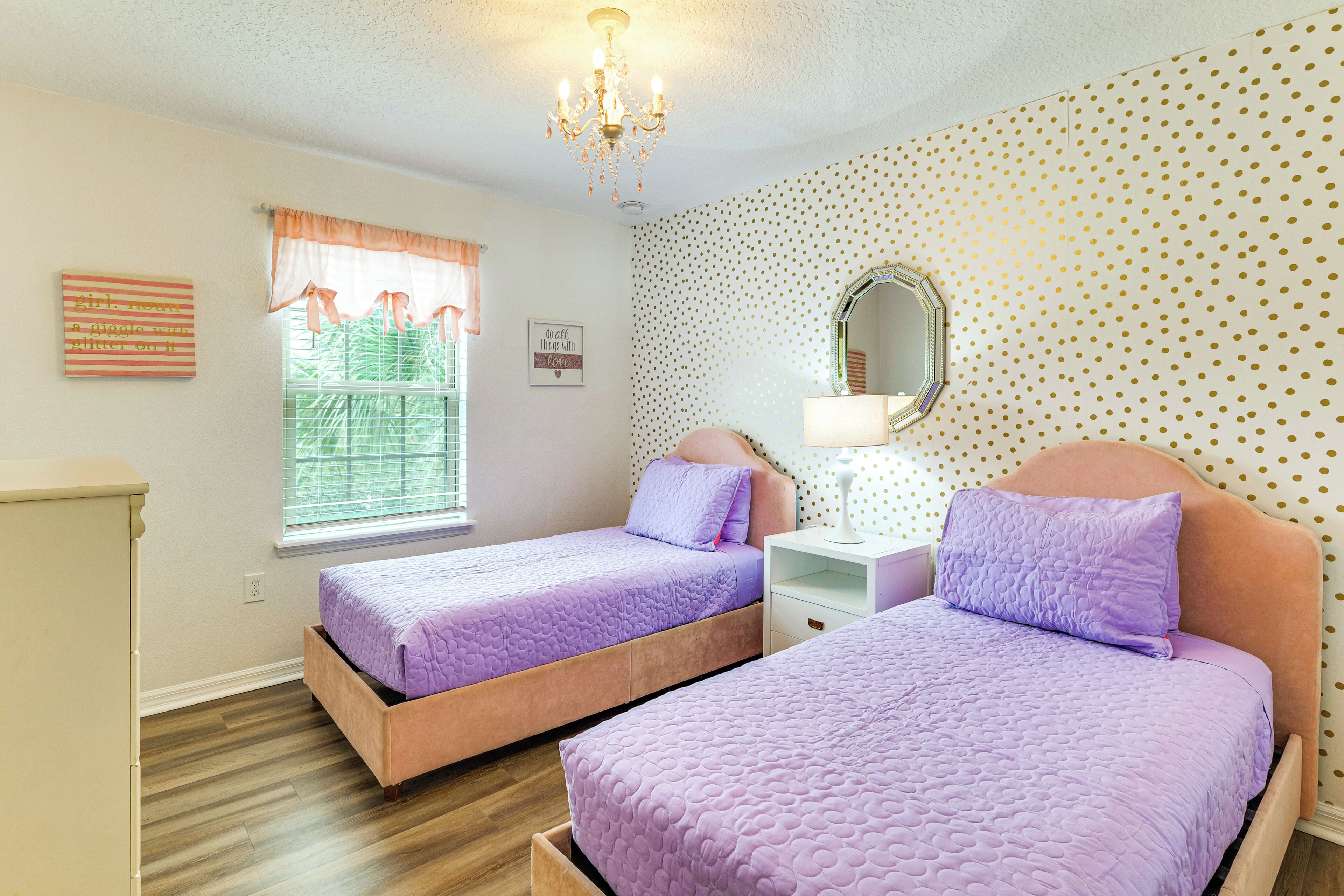 A bedroom with two beds, a dresser, a window with curtains, a chandelier, a mirror, and a wall with a polka dot pattern.