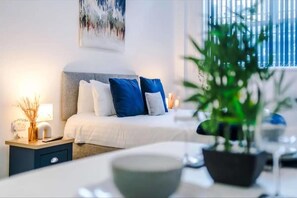 A modern bedroom with a bed, bedside table, lamp, and a potted plant. - Bolton City Oasis - Cozy Apartment Rental (Bolton)