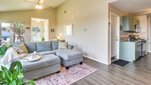 Ein Wohnzimmer mit grauem Ecksofa, Deckenventilator und einer Küchenzeile mit türkisfarbenen Schränken und einer Mikrowelle.