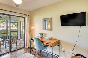 Quarto com escrivaninha de madeira, uma cadeira, televisão fixada na parede e uma porta de vidro deslizante para área externa.