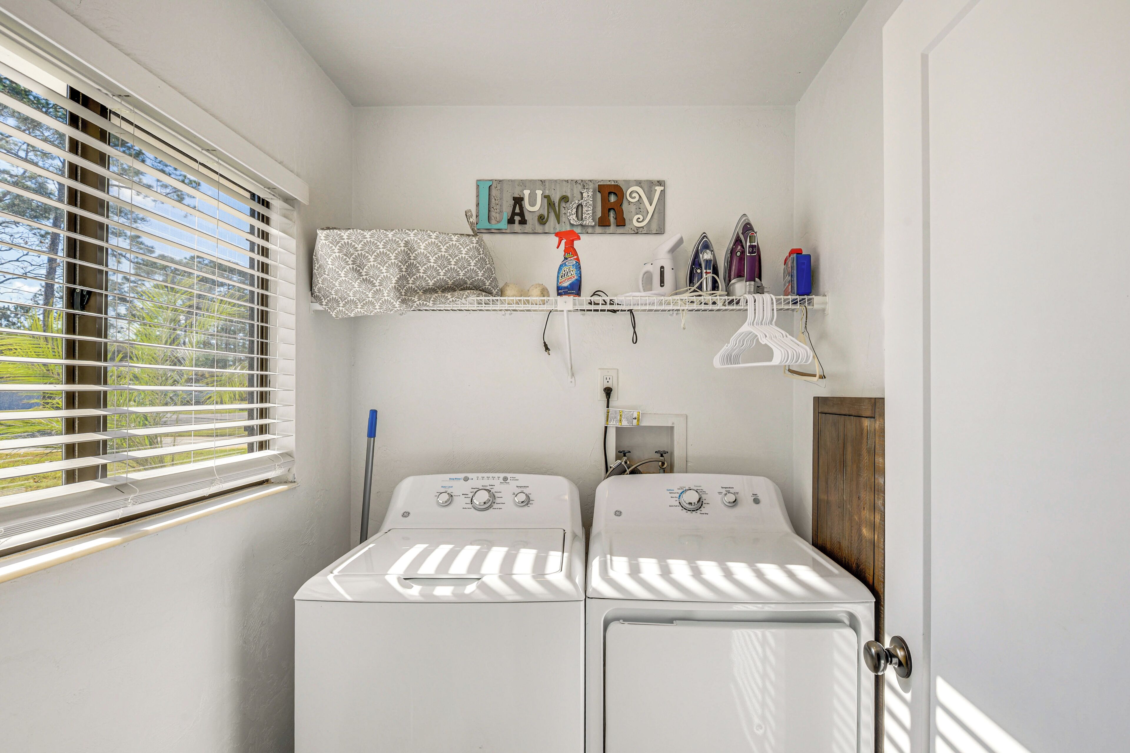 A laundry room with two washing machines, a shelf with laundry supplies, and a window with blinds.