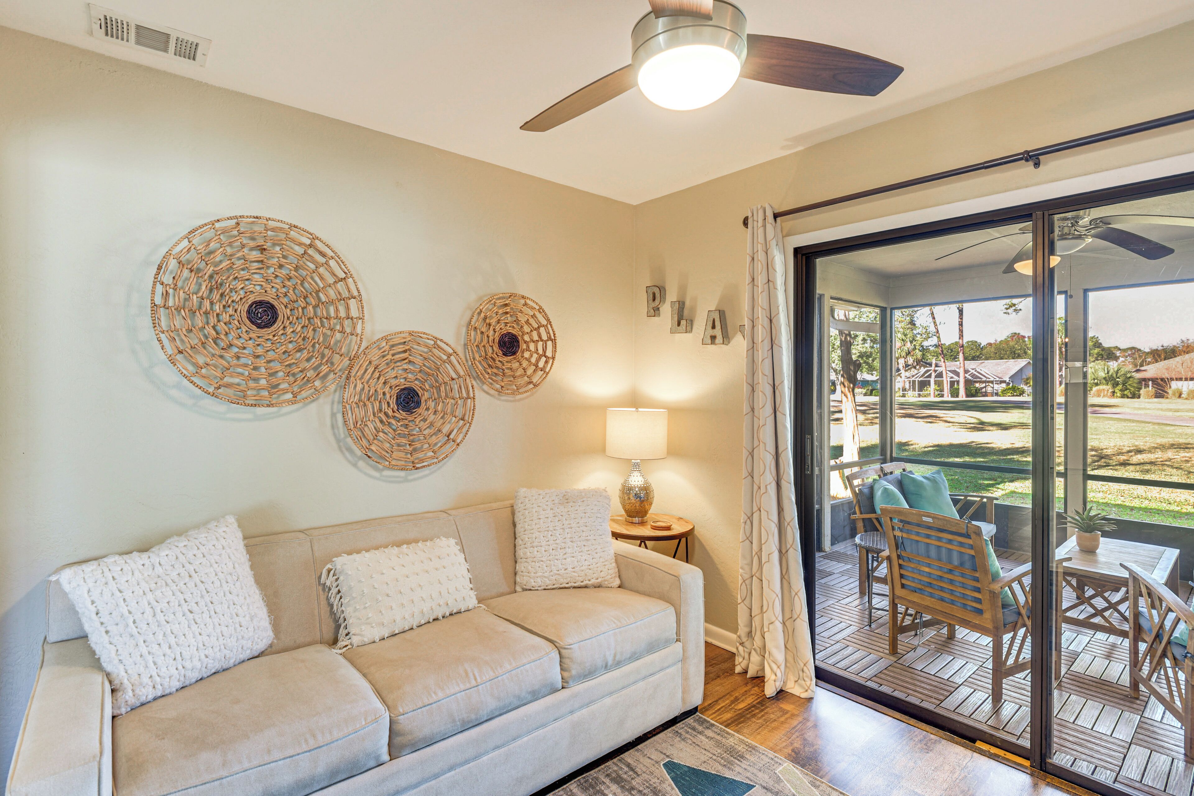 Un salon avec un canapé beige, un ventilateur de plafond et une vue sur une terrasse extérieure, accessible par une porte vitrée.
