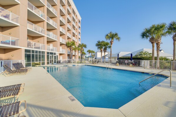 A swimming pool with sun loungers and palm trees, surrounded by a multi-story building with balconies. - Ocean View, Walk to Beach: Gulf Shores Condo (Gulf Shores)