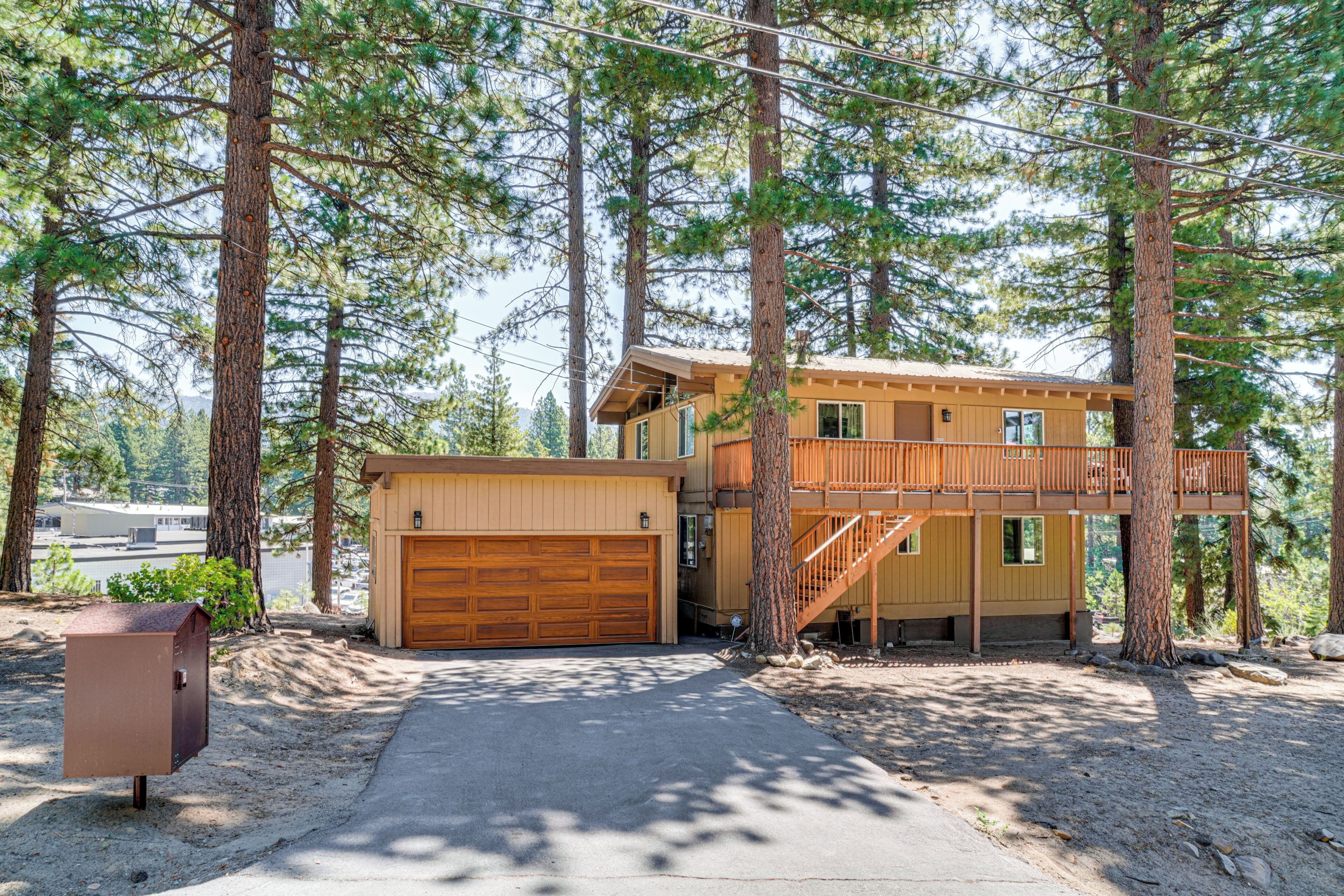 A two-story cabin with a wooden deck and a garage, surrounded by tall pine trees.