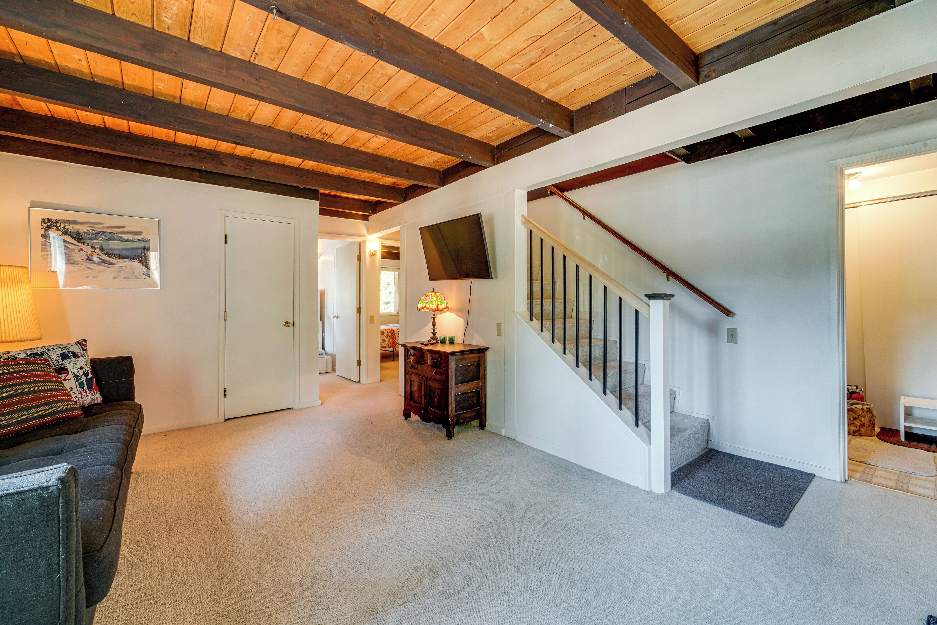 A spacious living area with a wooden ceiling, a staircase, a flat-screen TV mounted on the wall, and a cozy seating area with a lamp.