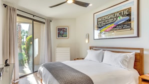 Een slaapkamer met een groot bed, een plafondventilator, een glazen deur naar een balkon en een ingelijst poster aan de muur.