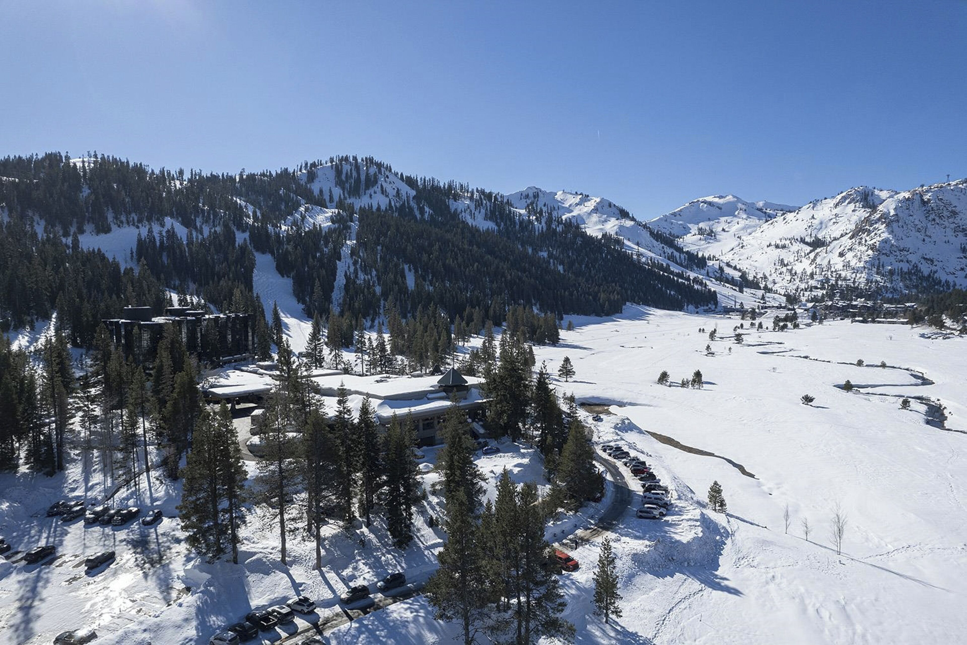 Une zone montagneuse enneigée, avec un bâtiment entouré d’arbres et des voitures garées.