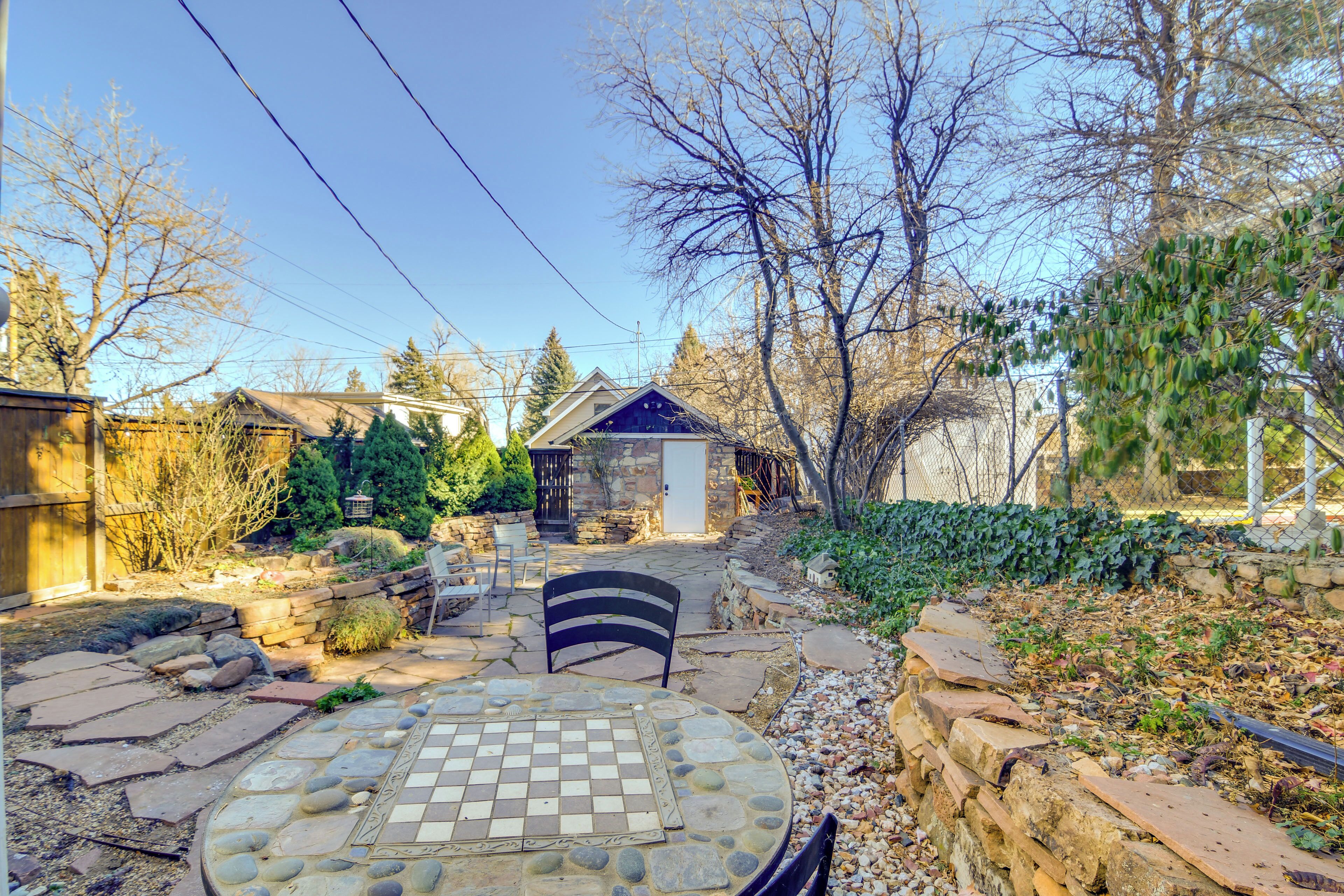A stone patio with a chessboard table, surrounded by a garden with various plants and a wooden fence.