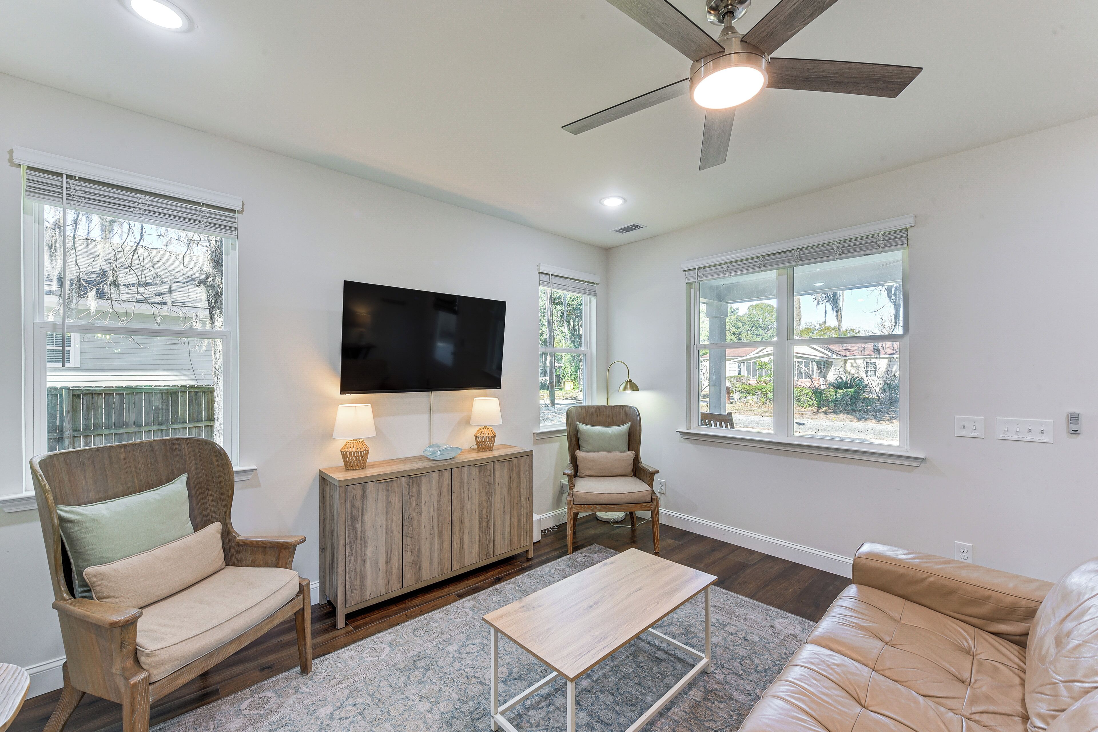 A living room with a ceiling fan, a TV, wooden furniture, and large windows.