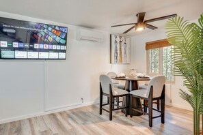 Zona pranzo con TV a parete che mostra servizi di streaming, ventilatore a soffitto e un'ampia finestra con persiane.