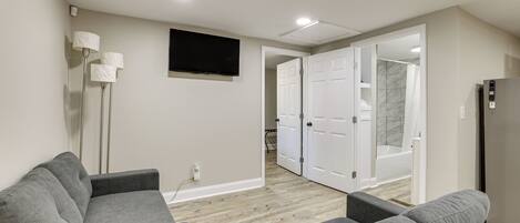 A modern living room with a grey sofa, a flat-screen TV, and a hallway leading to a bathroom with a shower and toilet.