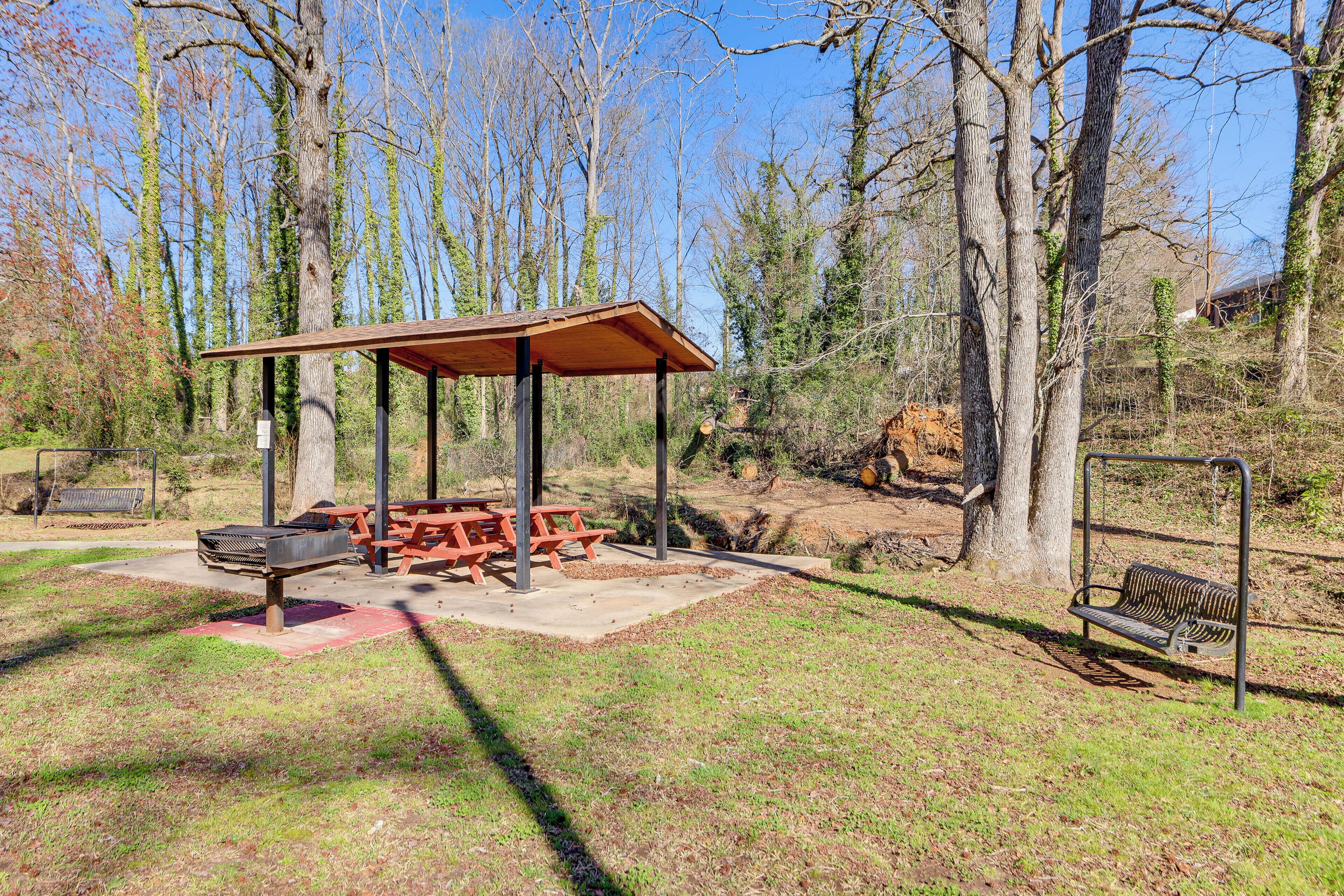 A covered picnic area with benches and a grill, surrounded by trees and a picnic table.