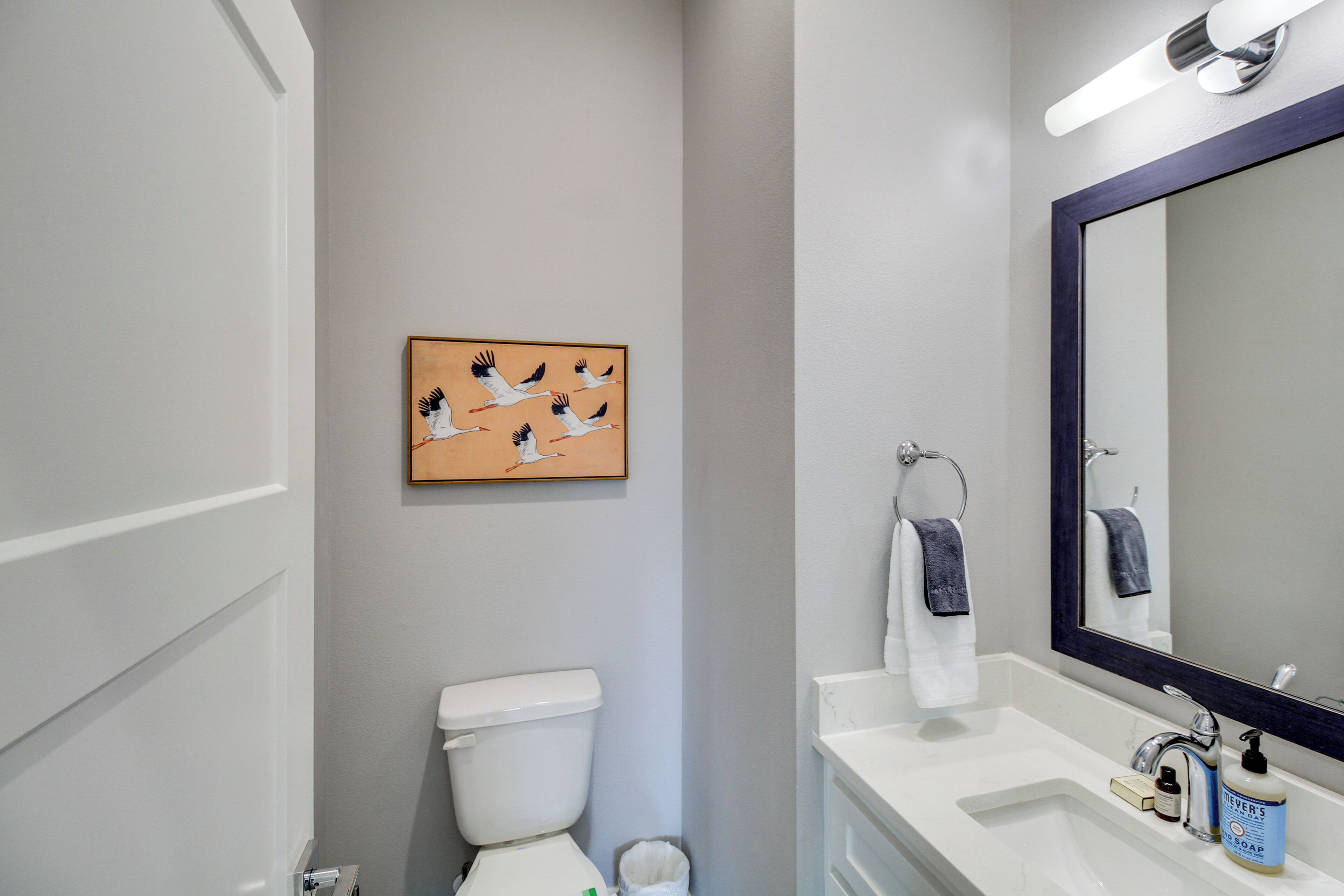 Ein Badezimmer mit Toilette, Waschbecken und Spiegel. Über der Toilette hängt ein gerahmtes Kunstwerk und eine Handtuch an der Wand.