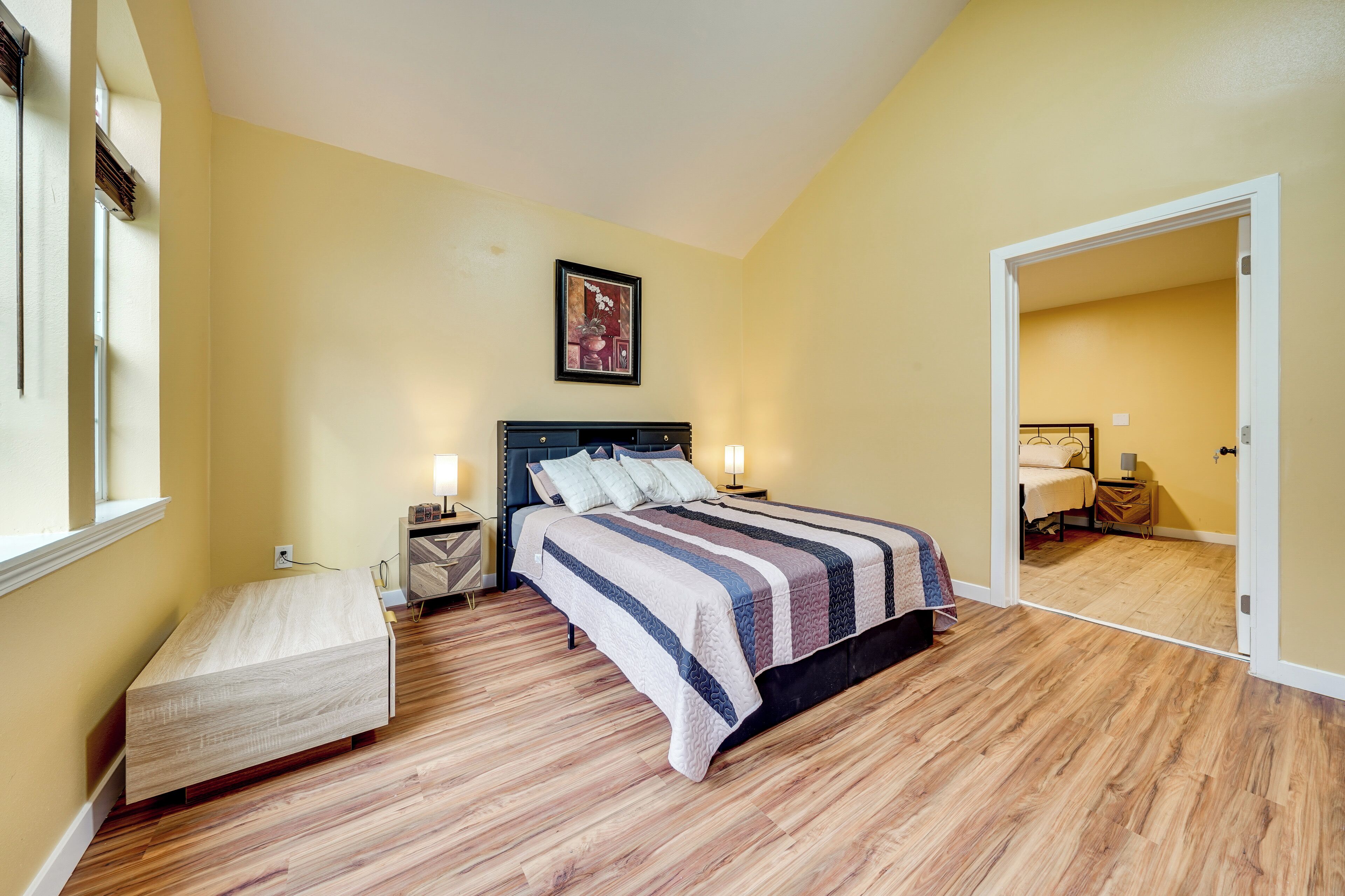 Une chambre à coucher comprenant un lit, des tables de chevet, une commode, un banc et une porte donnant sur une autre pièce.