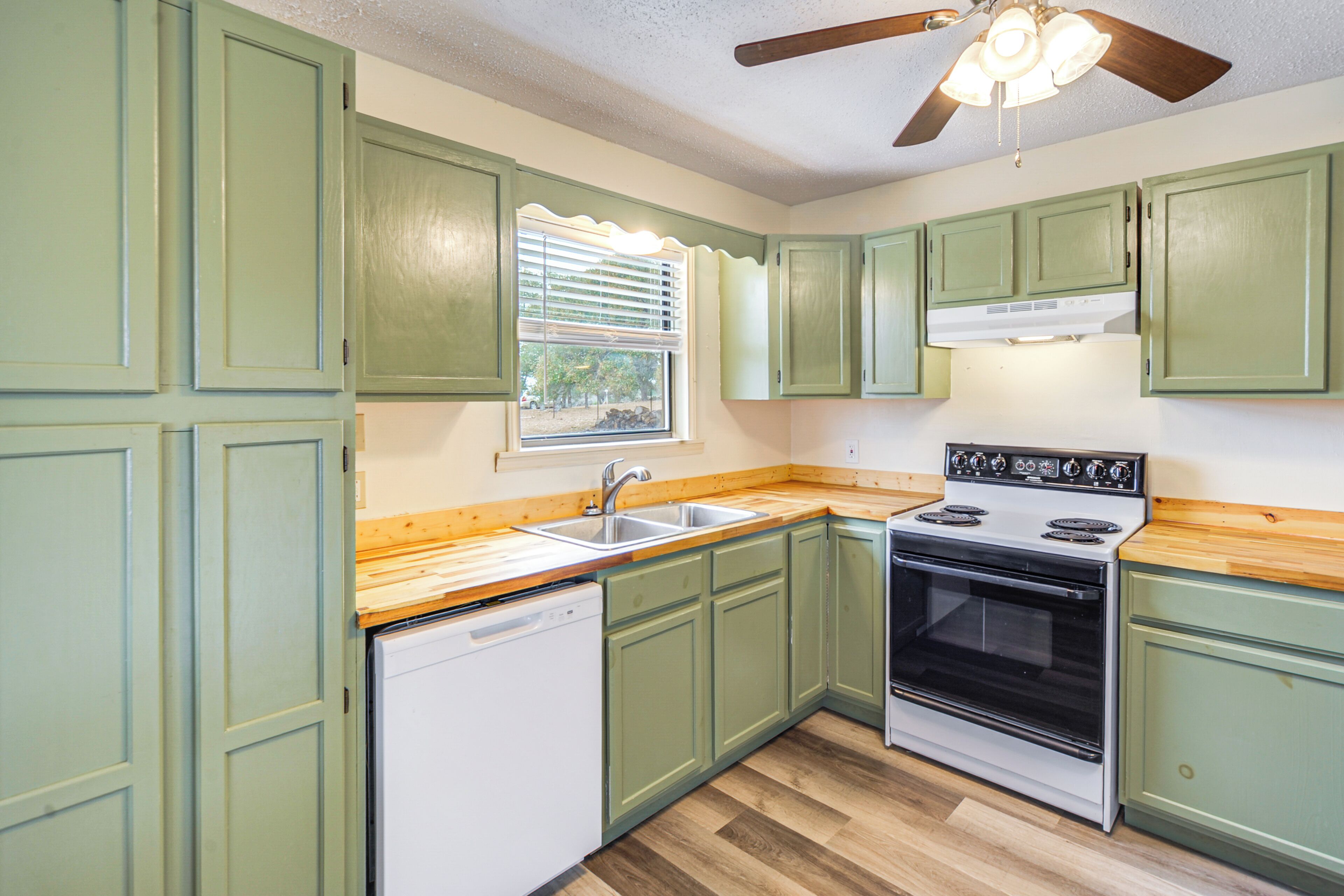 A kitchen with green cabinets, a white dishwasher, a stove, and a window with blinds.