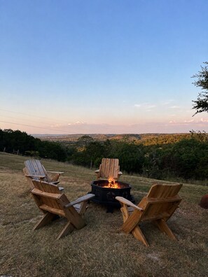 Una fogata con dos sillas Adirondack de madera orientadas hacia ella, ubicada en una colina cubierta de césped con vista a un paisaje lejano.