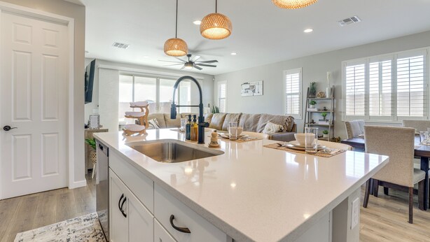 A modern kitchen with a large island, white cabinetry, and a built-in sink. There are wicker pendant lights, a ceiling fan, and a view of the living area with a sofa and dining table. - Serene Outdoor Space: Group Getaway in Maricopa! (Maricopa)