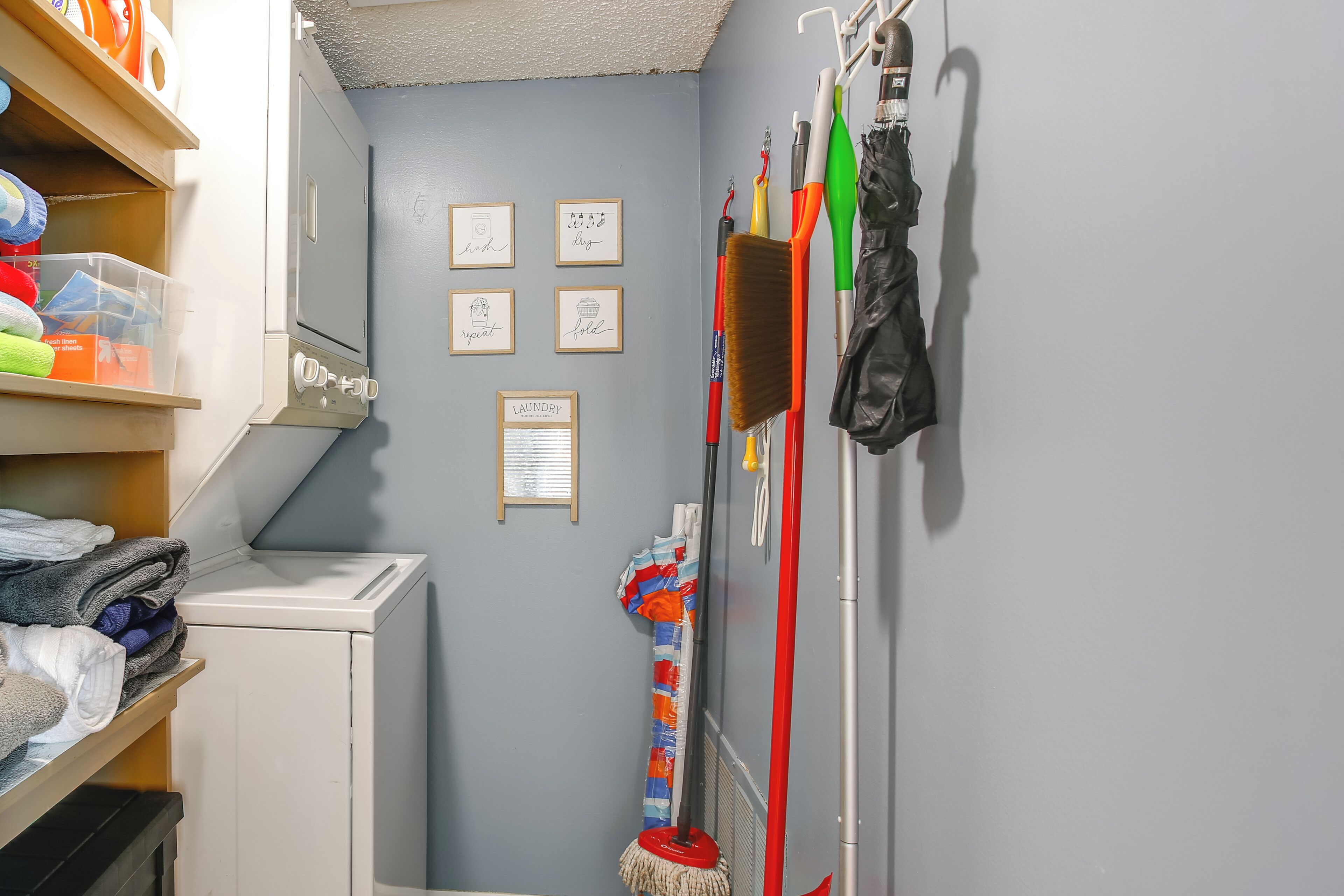 A laundry room with a washing machine, shelves with folded clothes, and various cleaning tools hanging on the wall.