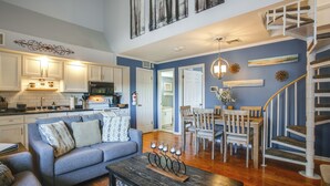 A spacious living area with a sofa, coffee table, and dining table. A kitchen with white cabinets and a staircase is visible. - Steps to Brigantine Beach: New Jersey Beach Condo (Brigantine)