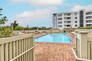 A swimming pool with a brick deck, surrounded by lounge chairs and umbrellas, with a multi-story building in the background. - Steps to Brigantine Beach: New Jersey Beach Condo (Brigantine)
