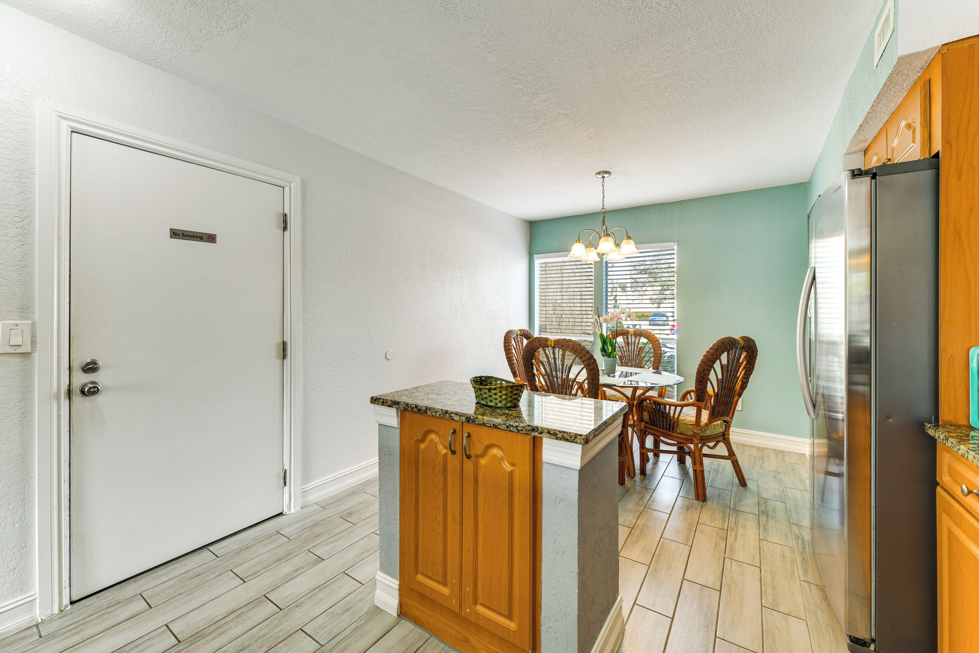 A kitchen with wooden cabinets, a stainless steel refrigerator, a dining area with wicker chairs, and a window with blinds.