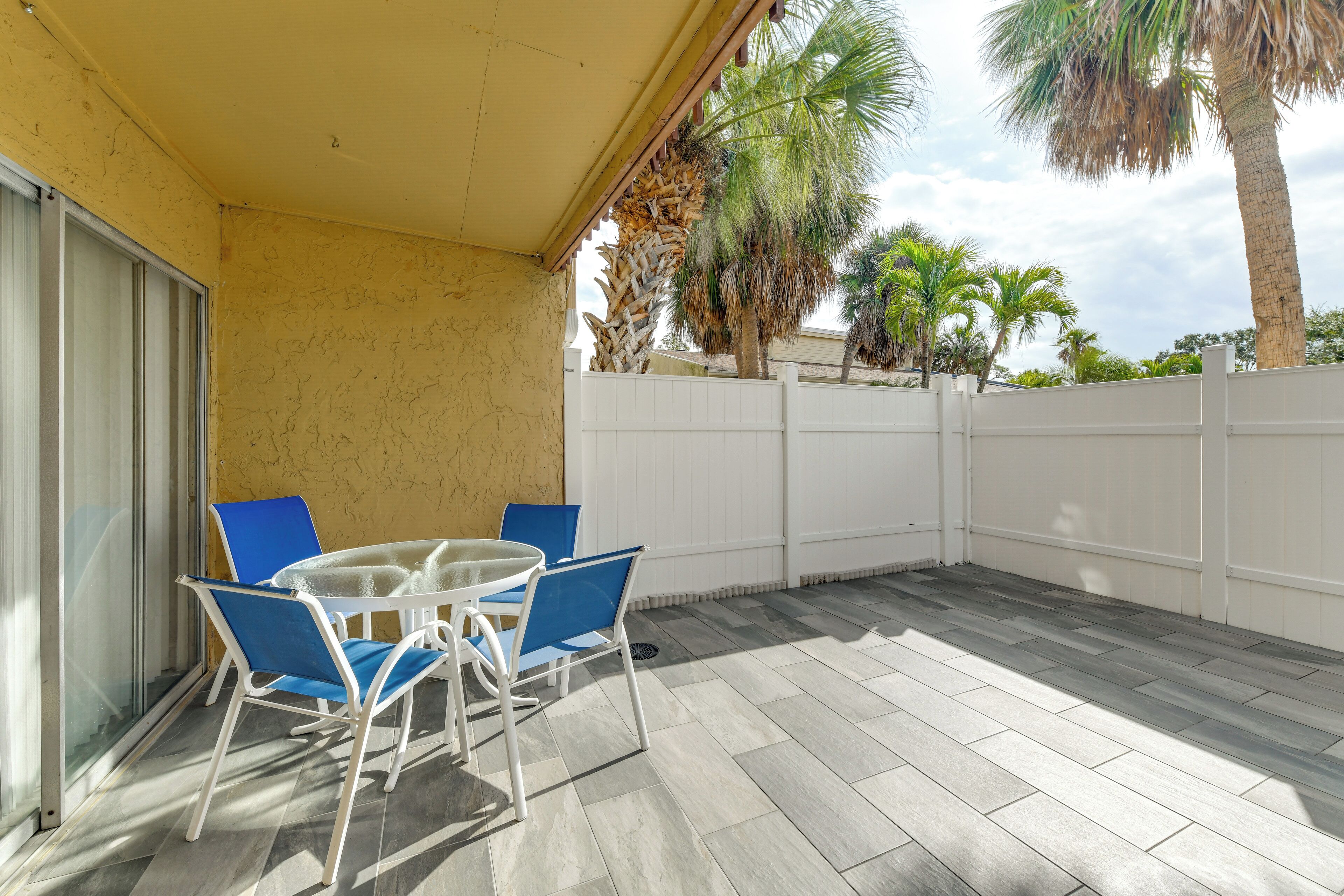 A covered patio with a table and chairs, a white fence, and palm trees in the background.