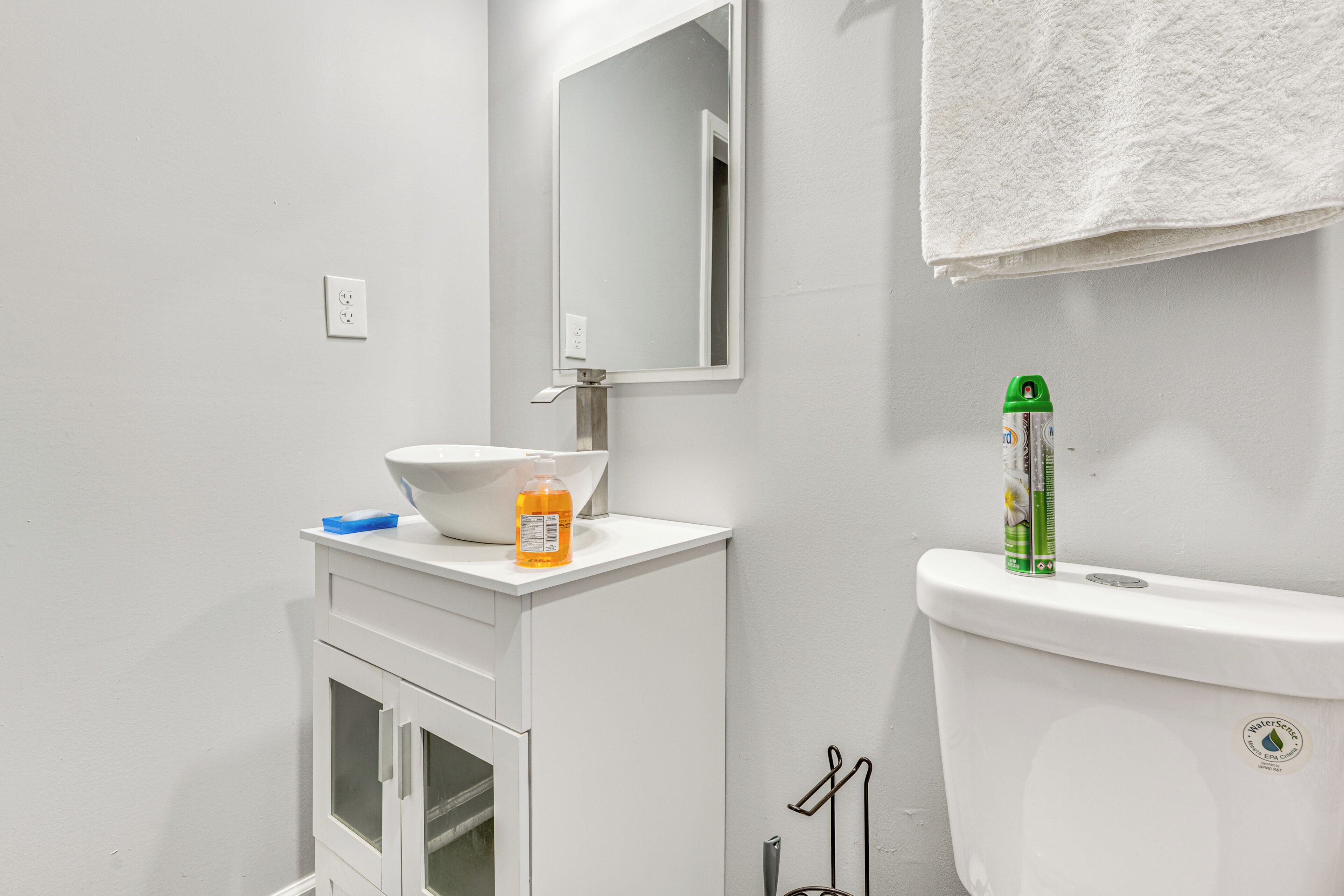 Une salle de bain avec un lavabo blanc, un miroir et des toilettes. On peut voir deux bouteilles sur le lavabo et une serviette accrochée au mur.