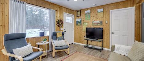 Un salón acogedor con paredes de madera, un ventilador de techo, un ventanal con cortinas, un televisor y dos sillones.