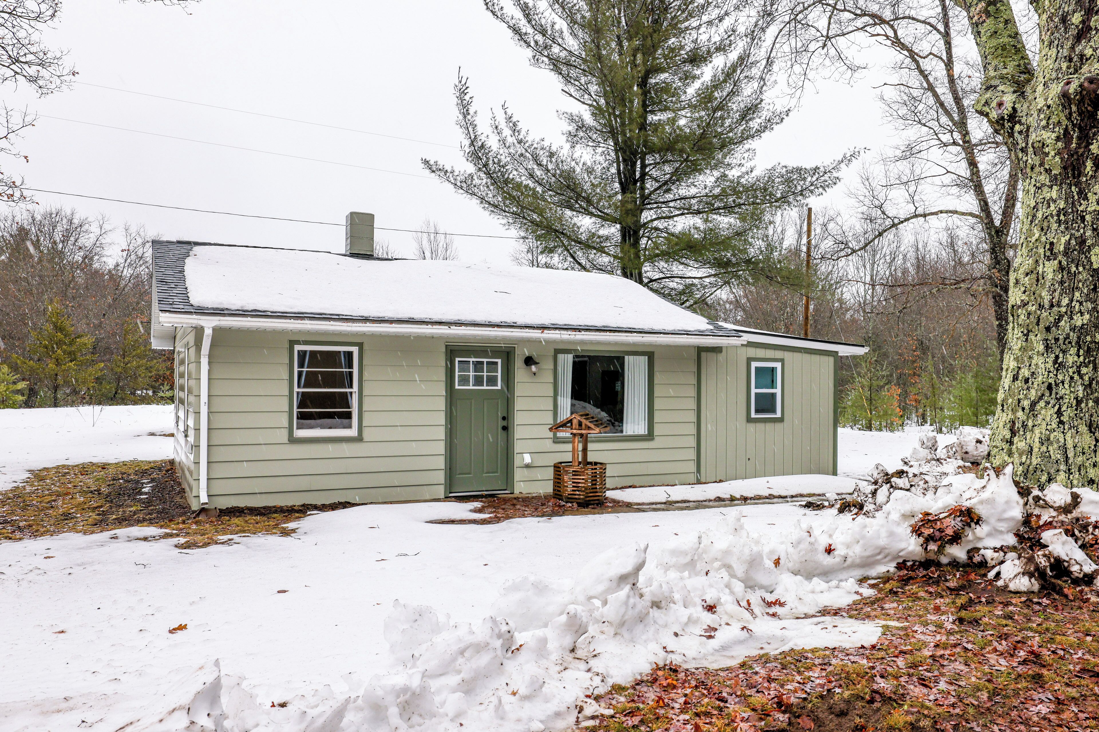 A small, single-story house with a green door and windows, surrounded by snow and trees.