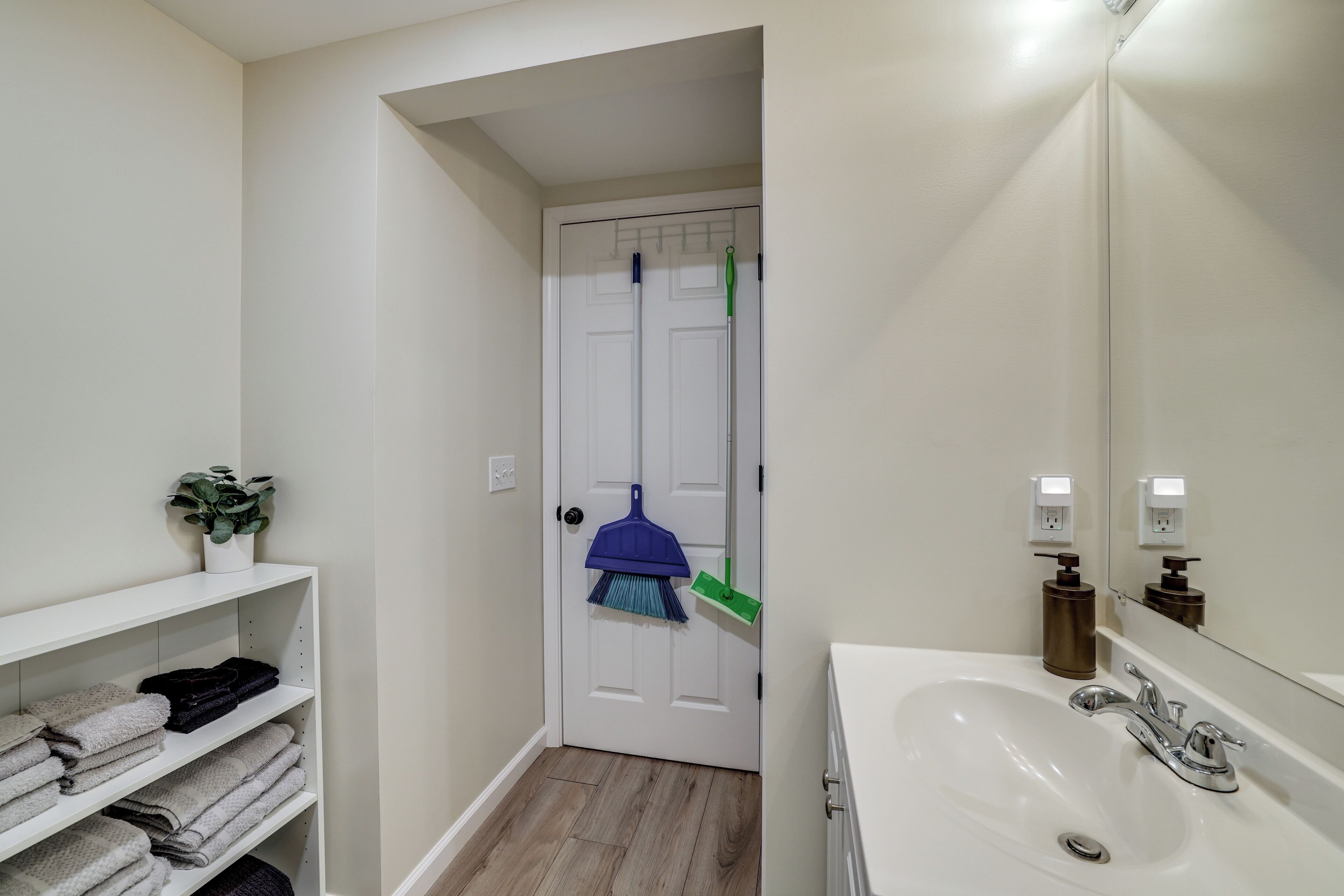 A bathroom with a white sink, a mirror, and a door with a broom and mop hanging on it.