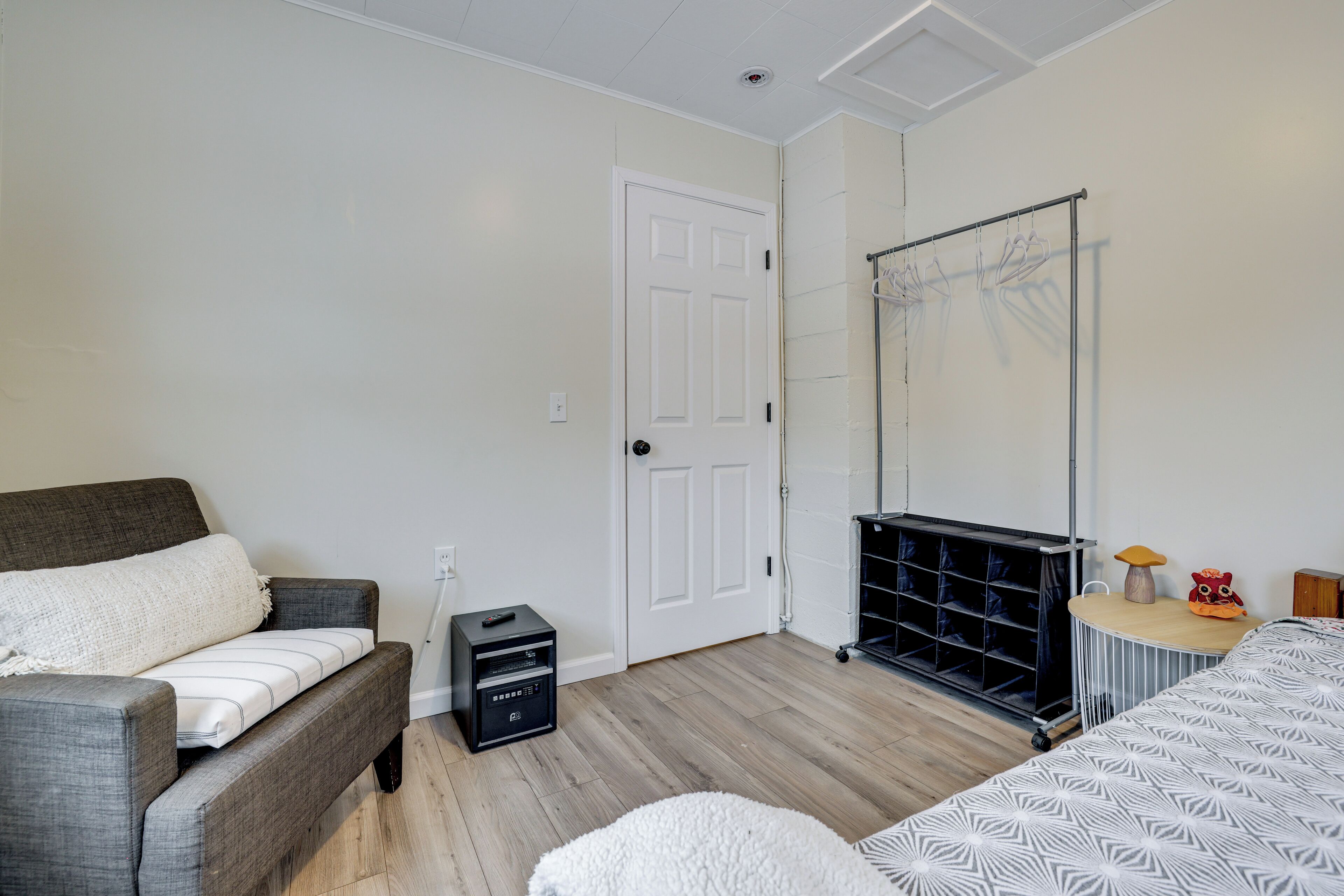 Een slaapkamer met een grijze bank, een bed met een bedspread met een patroon, een witte deur, een zwarte opbergunit en een kleine tafel met een lamp en decoratie.