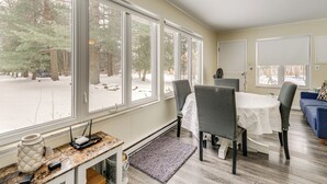 Un comedor con mesa y sillas, ventanas amplias con vista nevada y una pequeña mesa con un florero y dispositivos electrónicos.