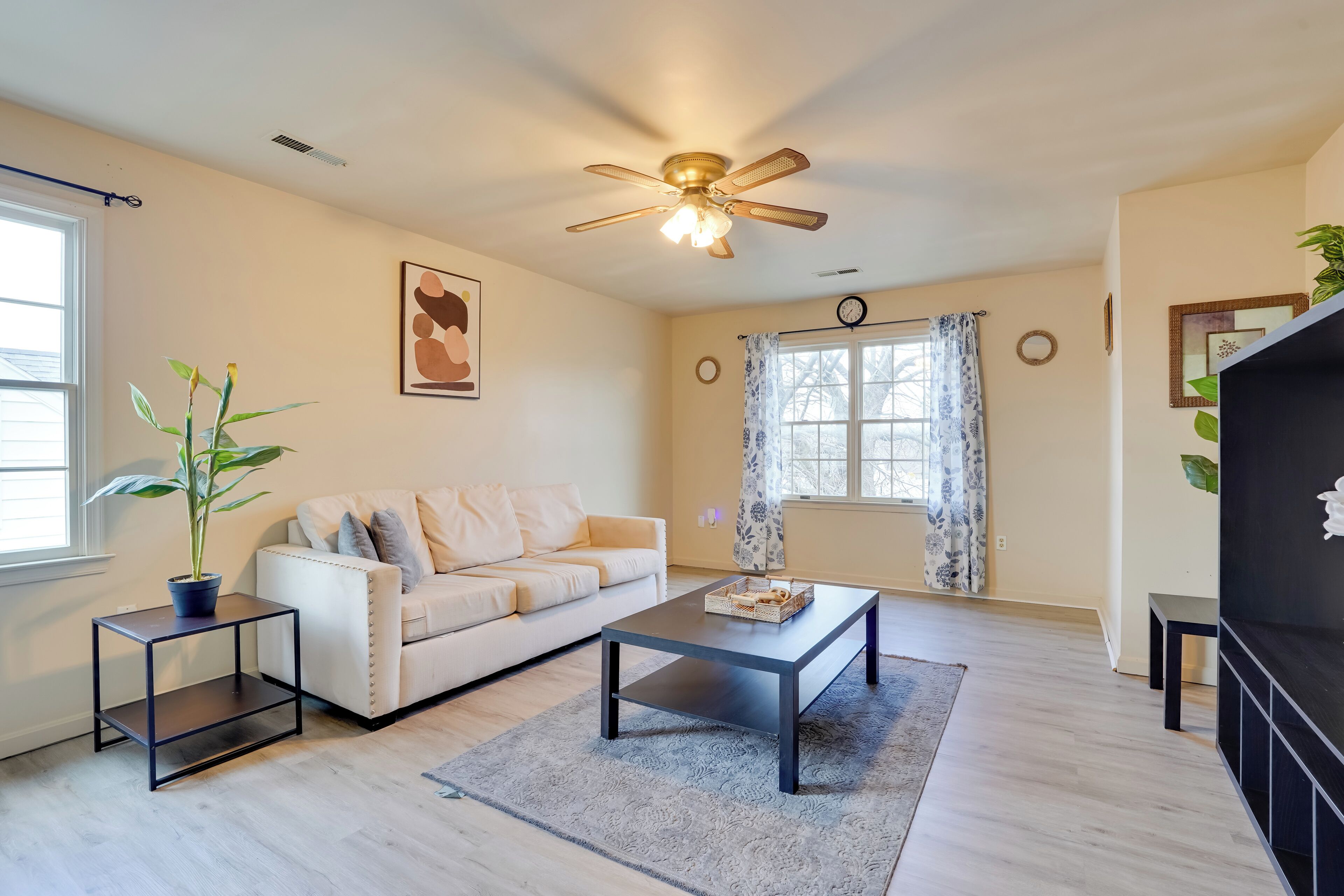 A living room with a white sofa, a coffee table, a ceiling fan, a potted plant, and a window with curtains.