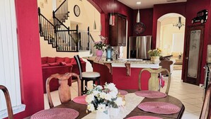 A dining area with a long table set for six, surrounded by chairs. A kitchen is visible in the background with modern appliances and a red accent wall. - SamAria - Two-story Villa (Deltona)